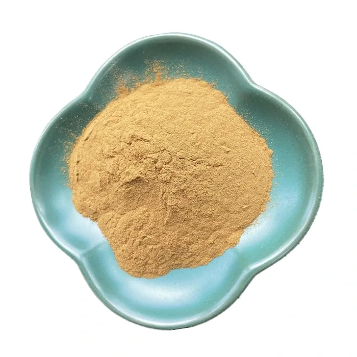 A wooden bowl of pale maca powder on a dark table beside dried roots, with green leaves behind